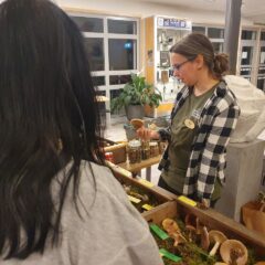 Nina håller en svamp i handen och står bakom ett bord med olika svampar i trälådor fyllda med mossa. I förgrunden två personer som tittar på svamparna.