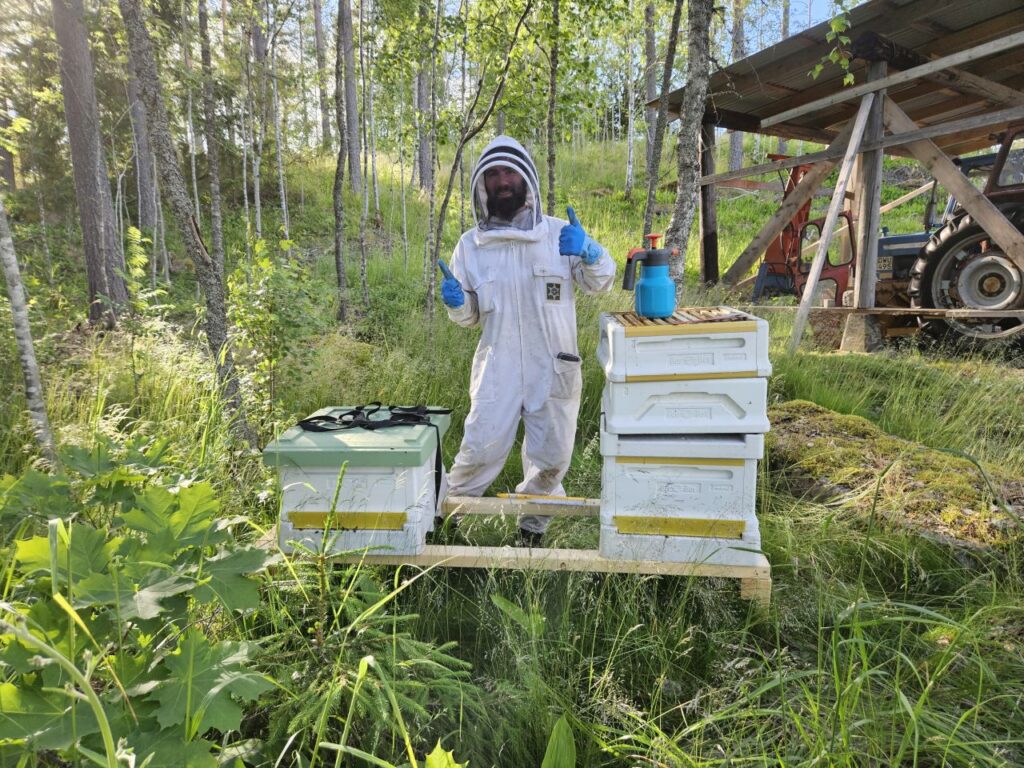 Kurser och undervisning 8 Thomas klädd i bidräckt står i en skogsmiljö framför två bikupor på en bänk. I bakgrunden en carport med en traktor. Solen skiner.