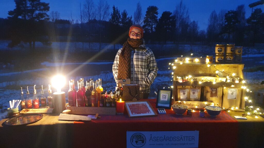 Foto på en person i vinterkläger med röd halsduk och mössa som står bakom ett bord med honungsglas, vinägerflaskor och paket med torkade svamp. Det är mörkt ute, bordet belyses med ljusslingor.